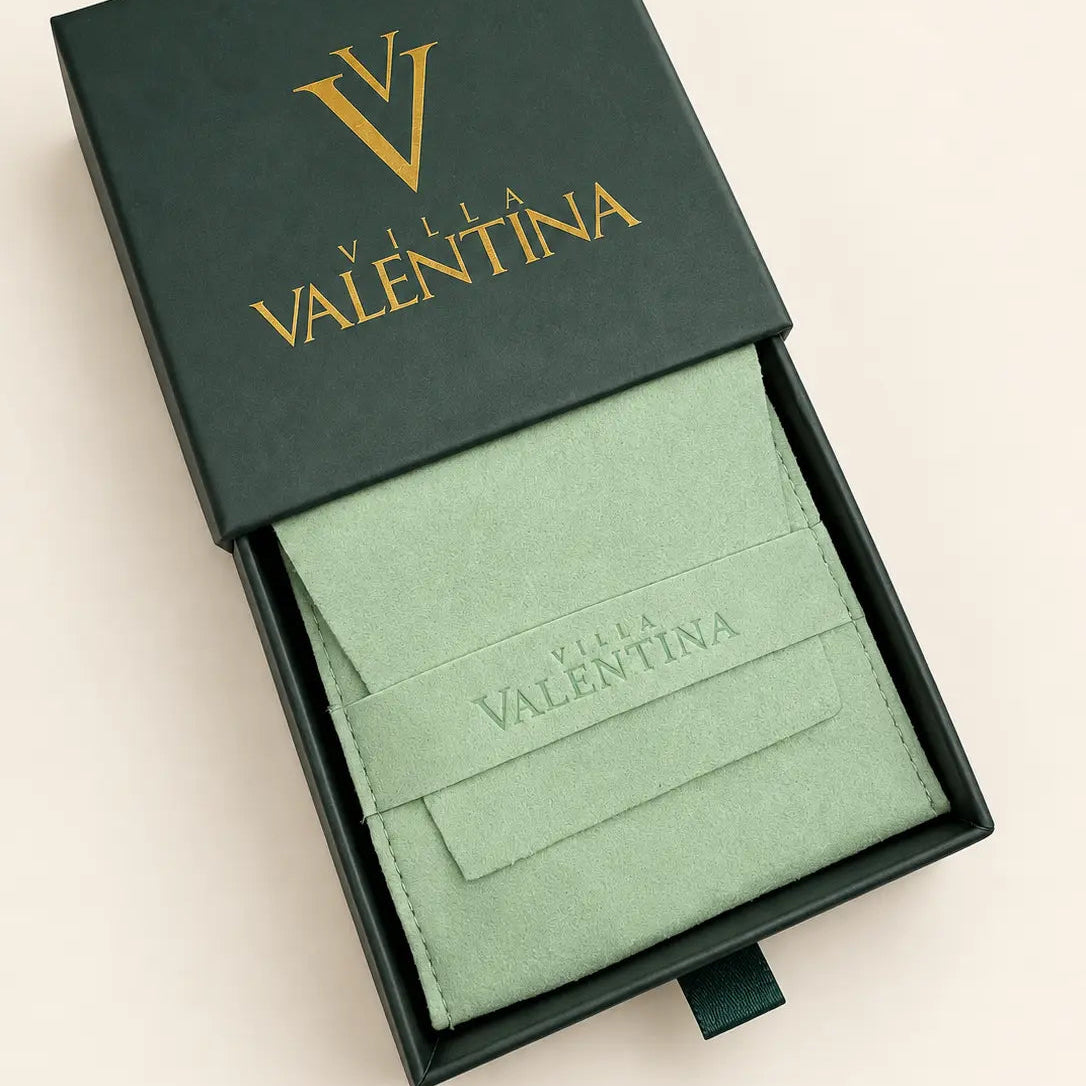 A dark green Valeria Valentina gift box, partially open, revealing a light sage green velvet pouch with the brand name debossed on it. The lid features a gold 'V' logo and 'VALENTINA' text, set against a solid cream background.