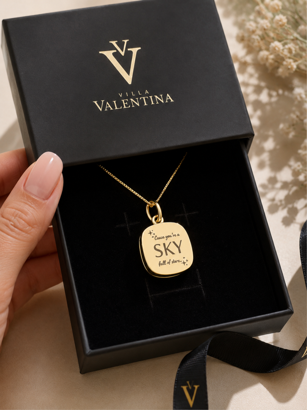 A gold pendant necklace with the engraved words 'I love you to SKY, built of dreams & stars' displayed inside an open black Villa Valentina gift box, held by a hand.