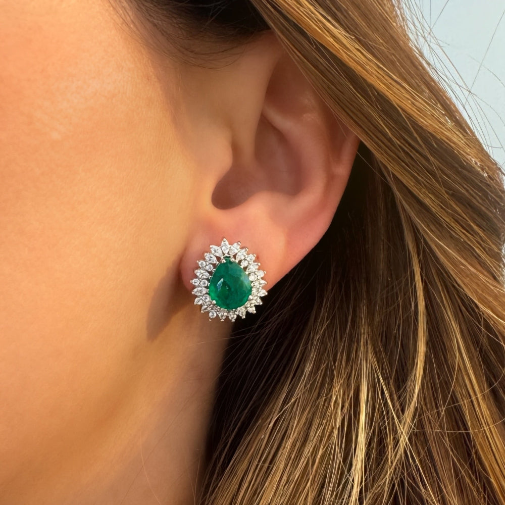 A close-up of a woman's ear wearing a teardrop-shaped emerald green gemstone stud earring surrounded by a halo of small clear stones in a silver setting.