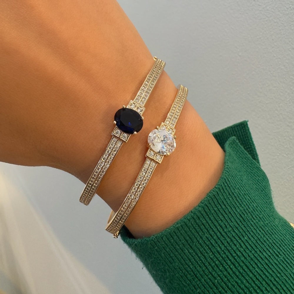 Two gold bracelets with gemstones on a wrist against a neutral background