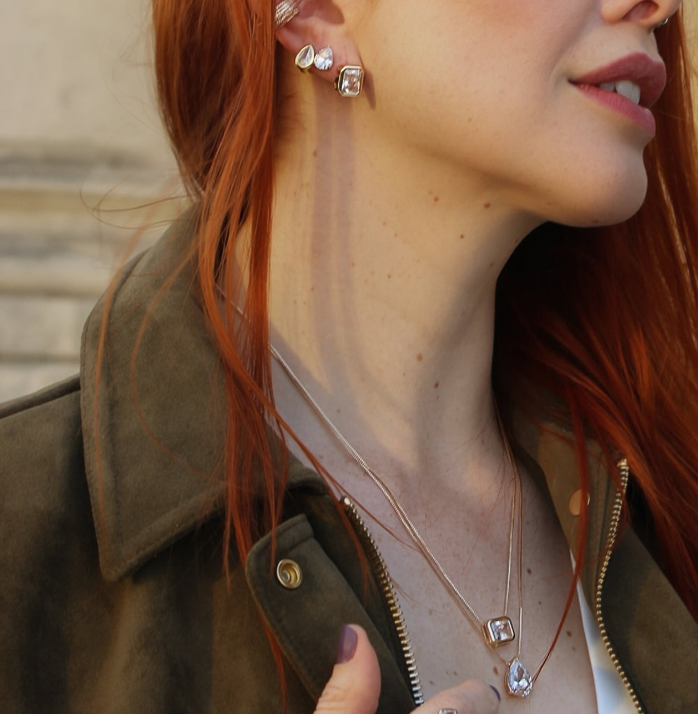 Close-up of a person with red hair wearing earrings and a necklace, with a blurred background.