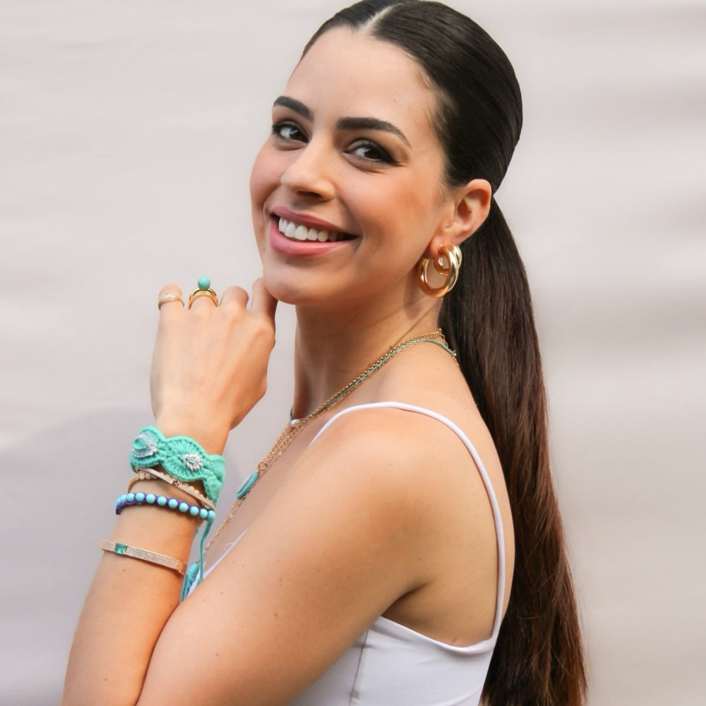A woman with dark hair in a ponytail, wearing a white camisole top and olive green pants, accessorized with turquoise and gold jewelry, posing against a light gray backdrop.