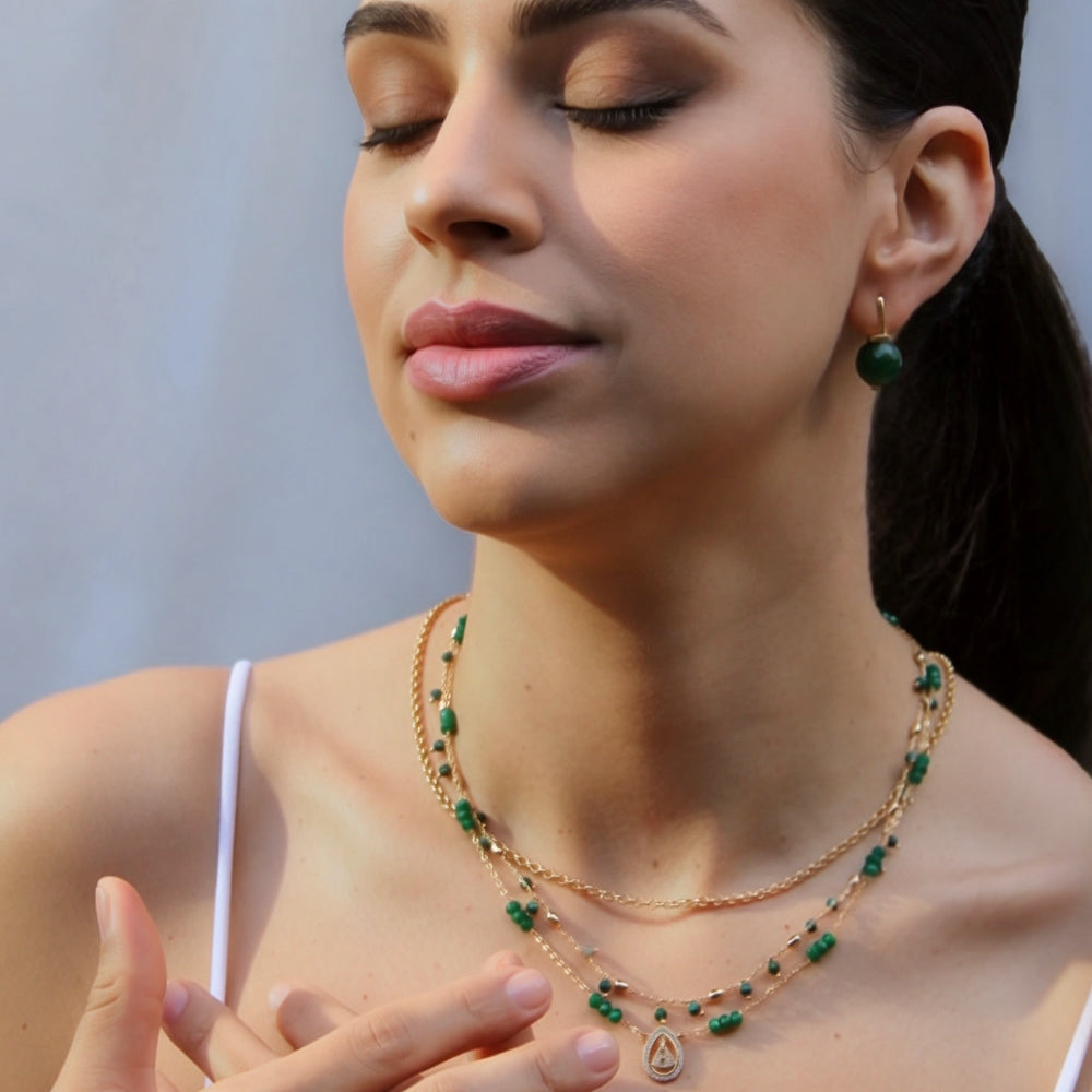 A woman with her eyes closed wears a layered gold necklace with green bead accents and matching green drop earrings, set against a soft gray background.