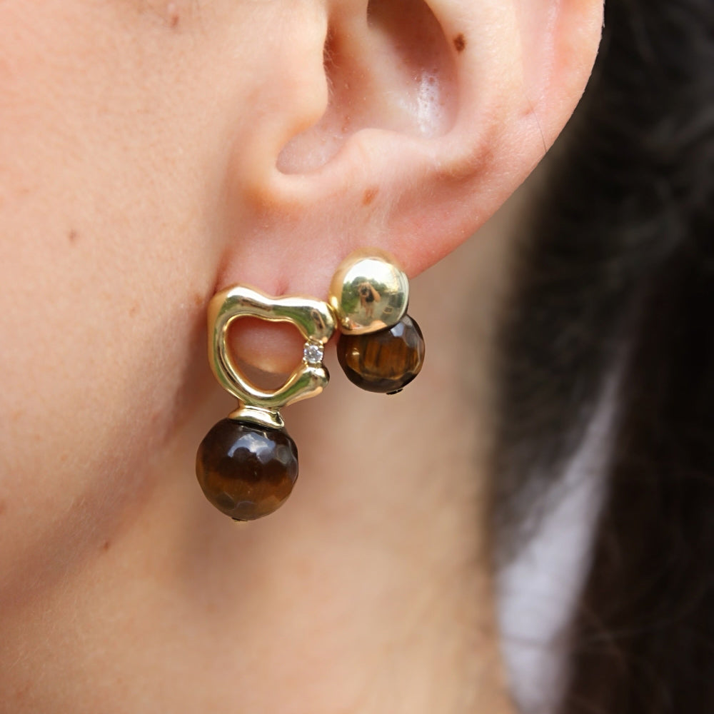 A close-up of a person's ear wearing a gold and brown drop earring with a heart-shaped gold stud, a small clear accent stone, and a larger round brown bead dangling below.