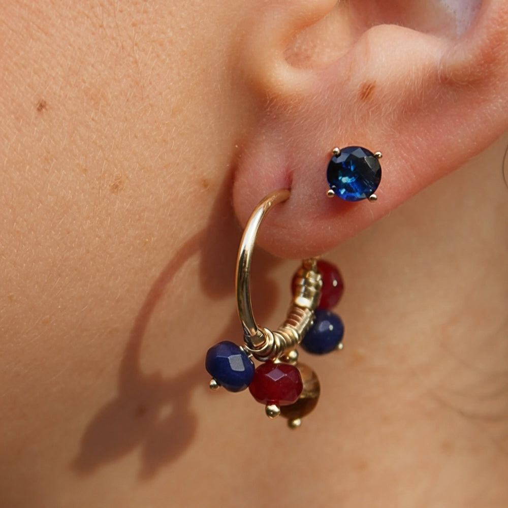 A close-up of a person's ear wearing a gold hoop earring adorned with blue and red faceted beads, alongside a small blue stud earring.