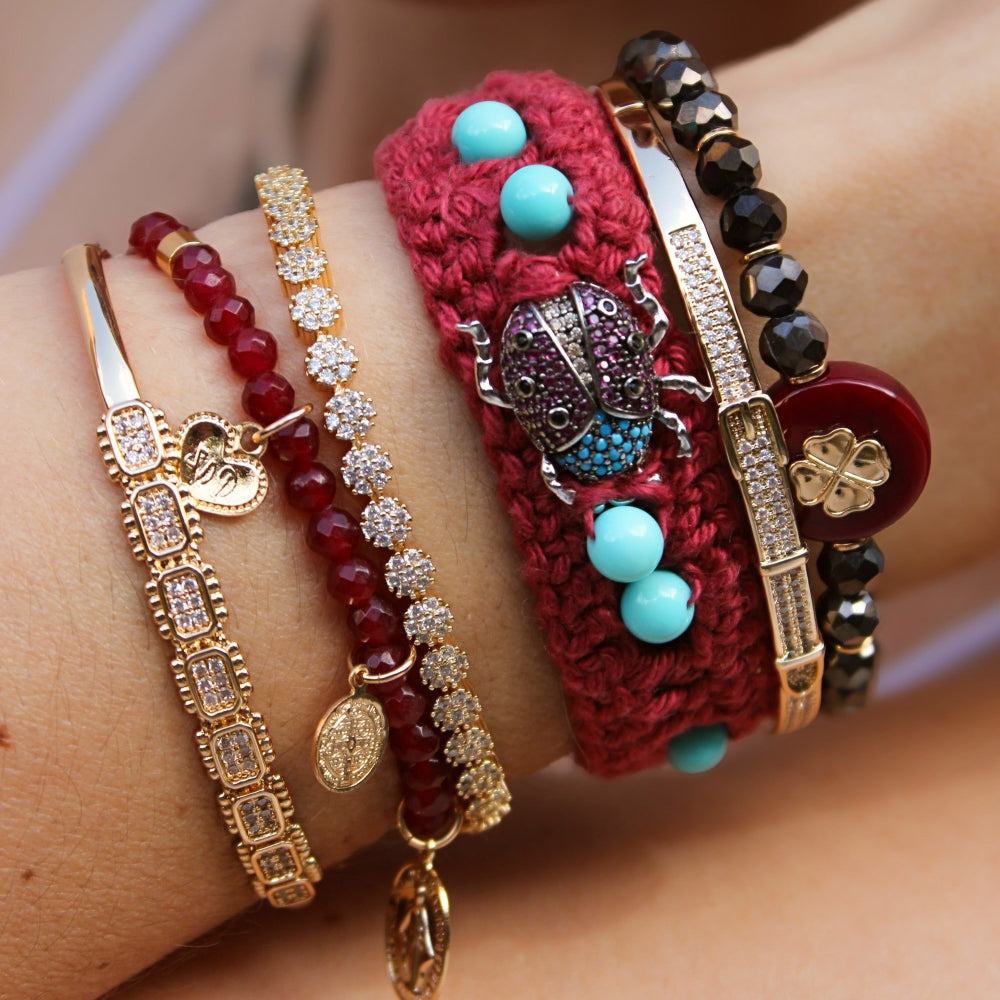 A close-up of a person's wrist wearing multiple bracelets, including a red crocheted bracelet with turquoise beads, a gold-tone beaded charm bracelet, and other mixed-material bracelets.