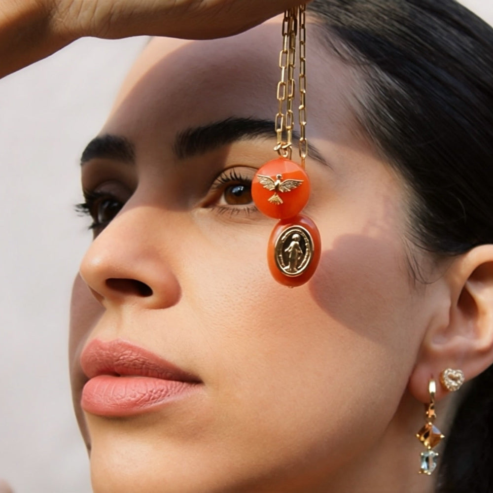 A close-up lifestyle shot of a model wearing a gold chain necklace with a large orange bead, a bee charm, and a circular medallion pendant, while holding the chain near her face.
