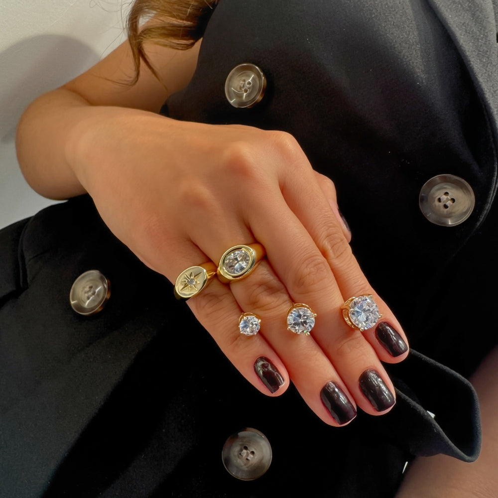 A close-up of a hand wearing multiple gold rings, including oval engraved rings and round bezel-set clear stones, styled against a black blazer.