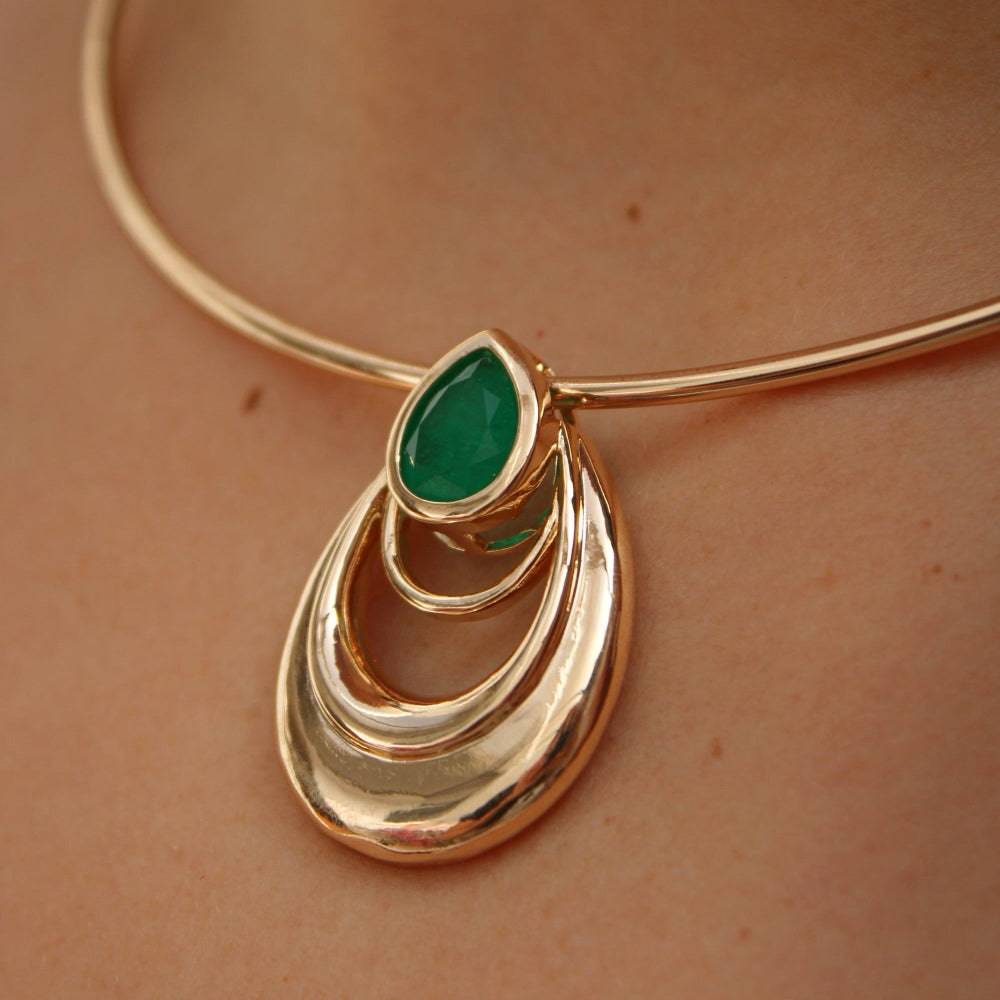 A close-up of a gold-toned choker necklace with a teardrop-shaped pendant featuring a green marquise gemstone, worn on a person's neck.