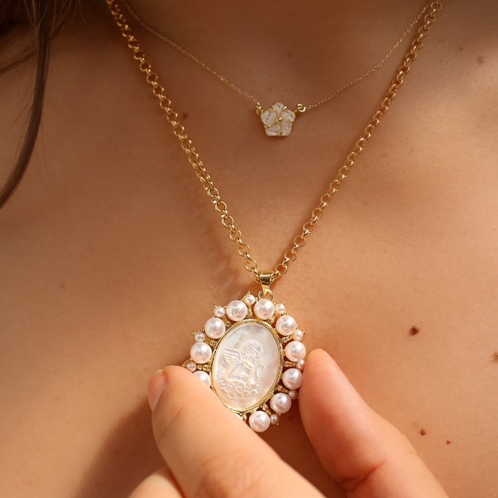 A close-up lifestyle shot of a model wearing a gold necklace with a white pearl-encrusted oval pendant and a matching white flower ring.