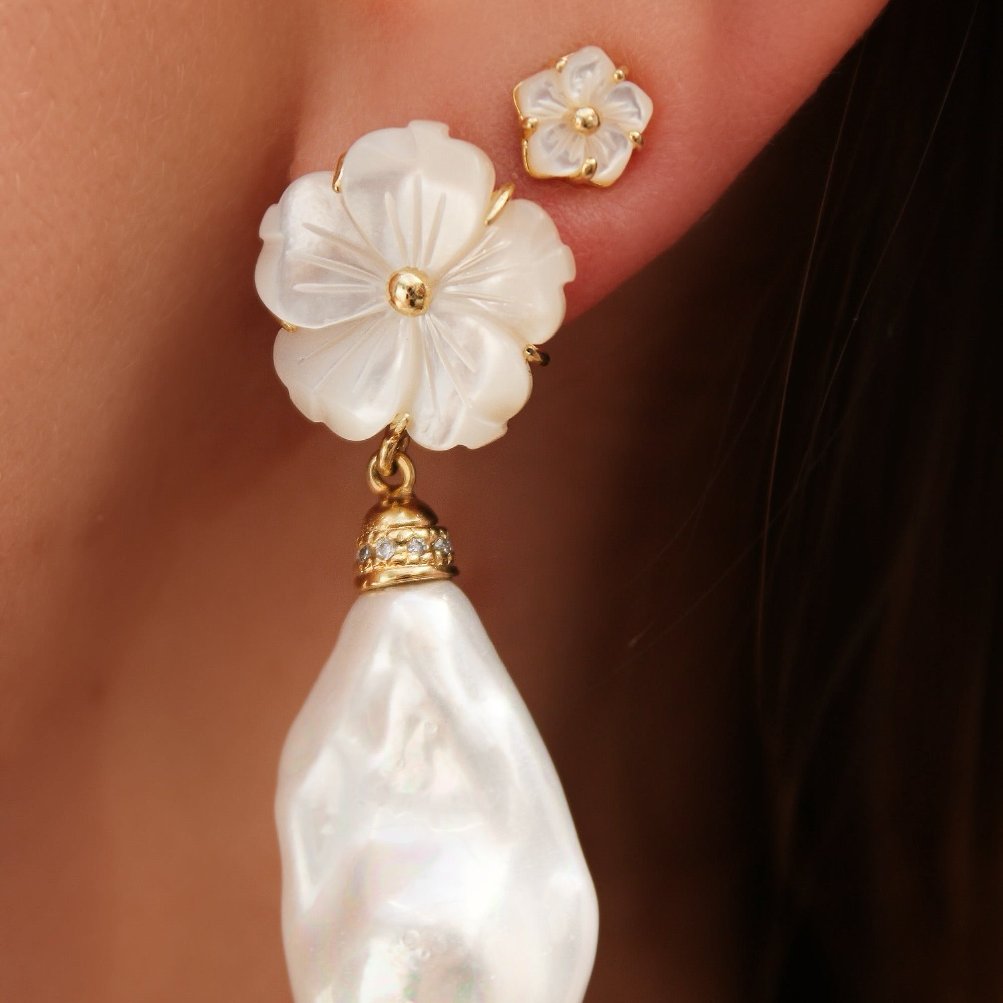 A close-up of a person's ear wearing a large white flower stud earring with a dangling baroque pearl and a smaller matching pearl stud on the lobe.