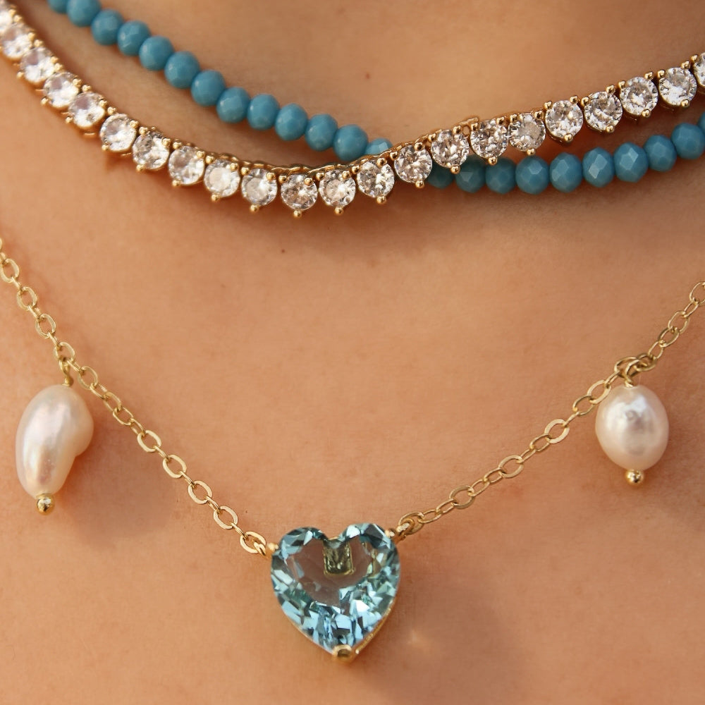 Close-up of layered necklaces worn on a person's neck, including a gold chain with a heart-shaped light blue gemstone pendant, a gold chain with white pearl beads, and a turquoise beaded necklace.