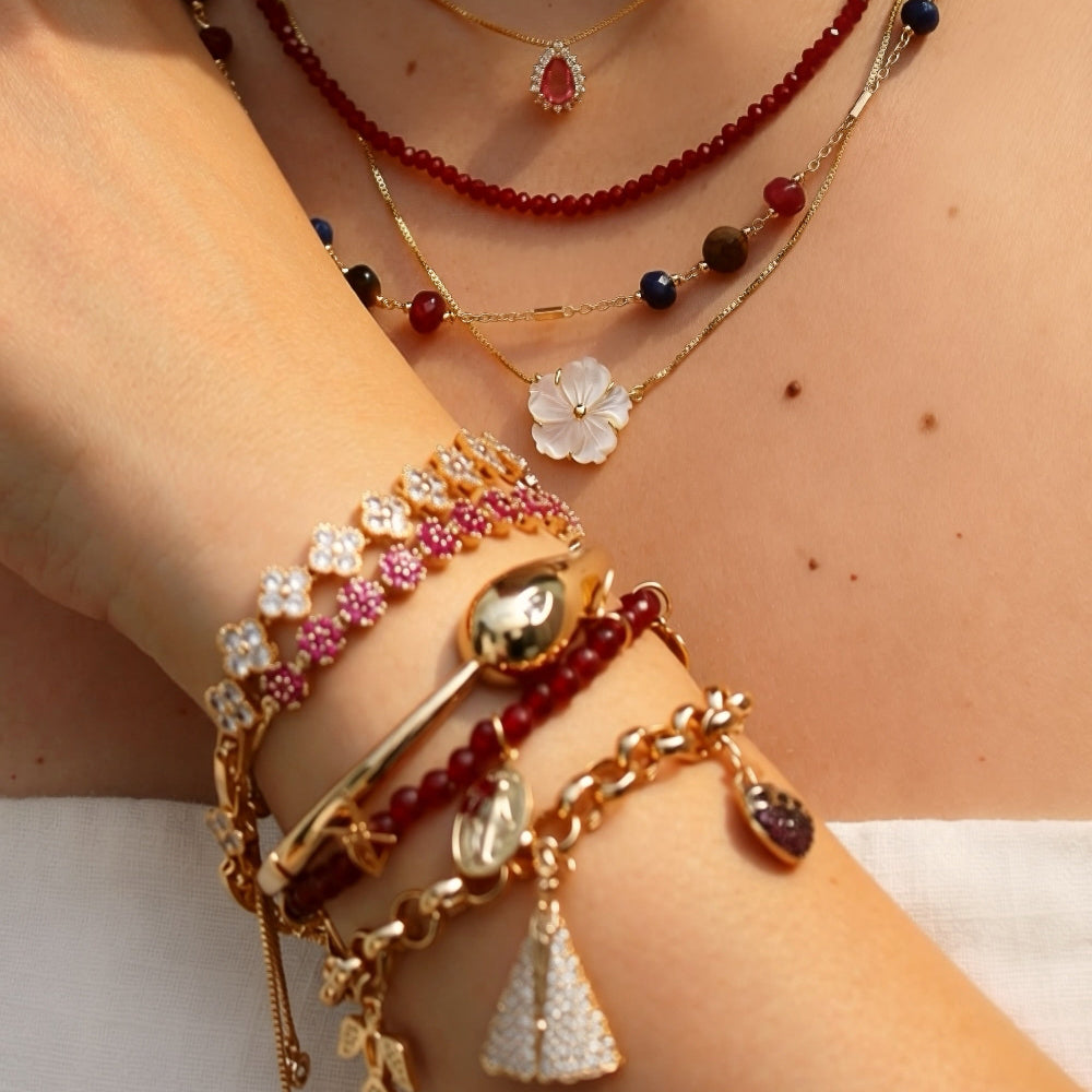 A close-up of a person's neck and wrist wearing multiple layered necklaces and bracelets. The jewelry includes gold-tone chains with floral charms, red beaded strands, clear rhinestone details, and dark bead accents.