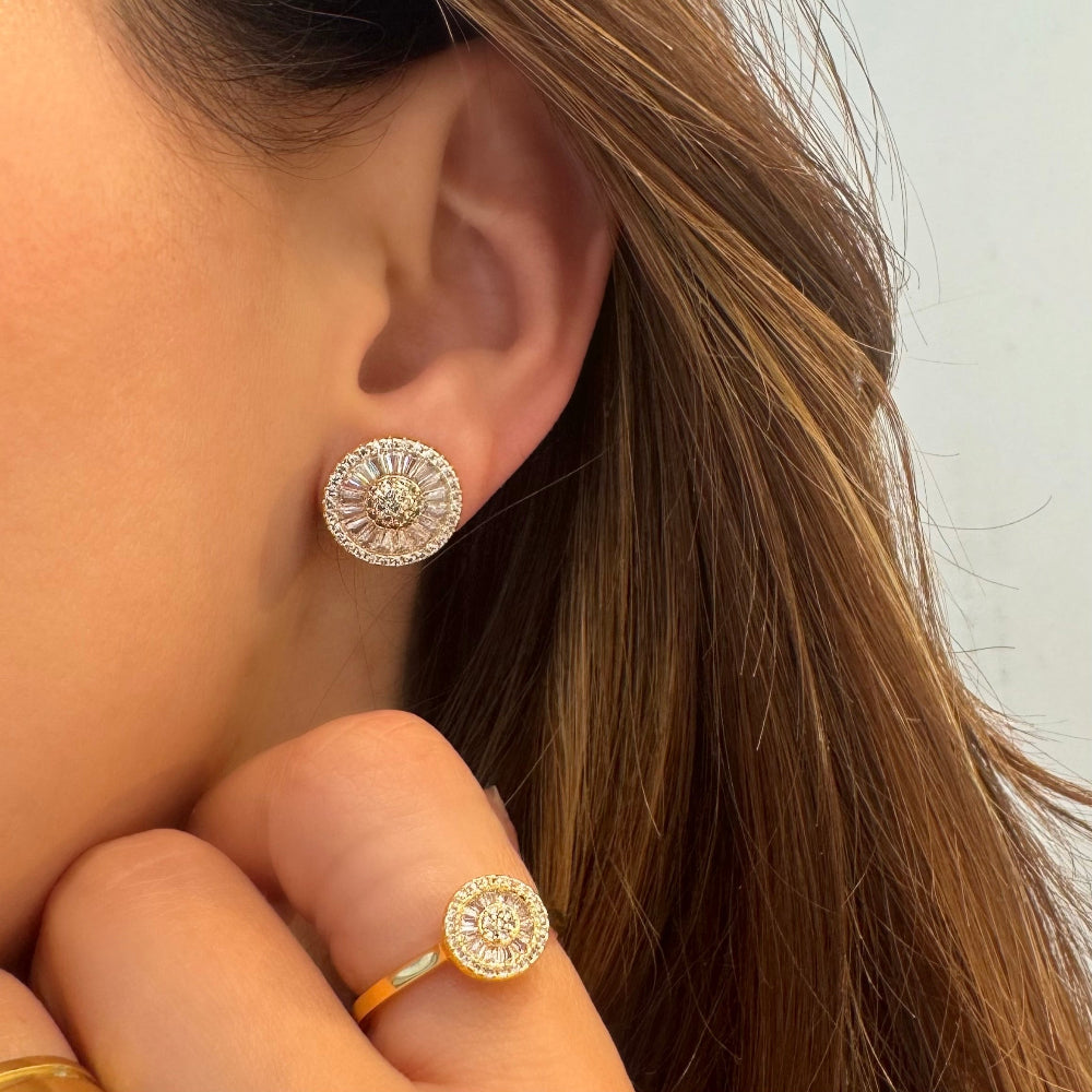 A close-up of a person's ear wearing a round silver-toned stud earring with a sunburst pattern of small clear stones, with a matching gold-toned ring also visible on the finger.