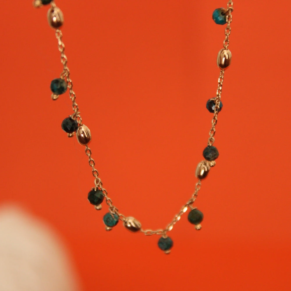 A delicate gold chain necklace with small dark green faceted beads and polished gold oval accents, shown against a vibrant orange background.