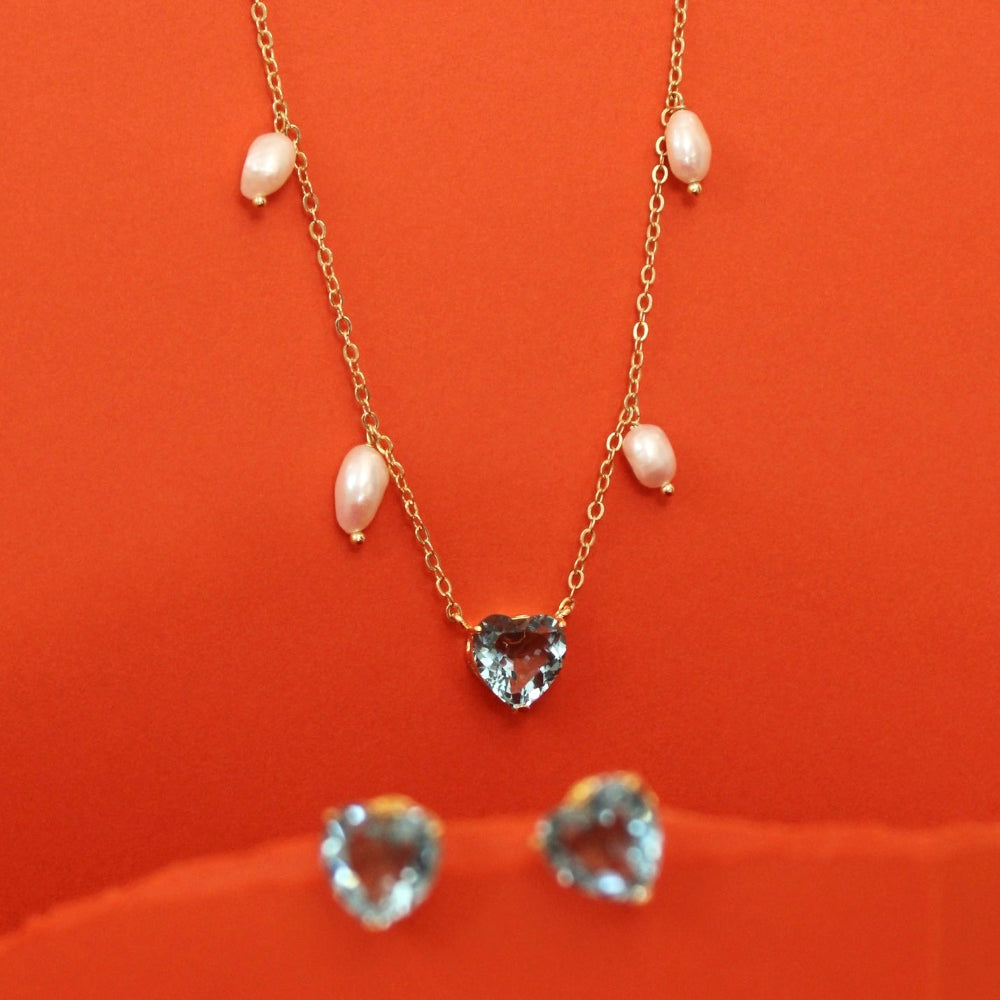A gold necklace with five small fresh water pearls and a heart-shaped clear gemstone pendant, displayed on a bright orange background with matching heart-shaped stud earrings below.