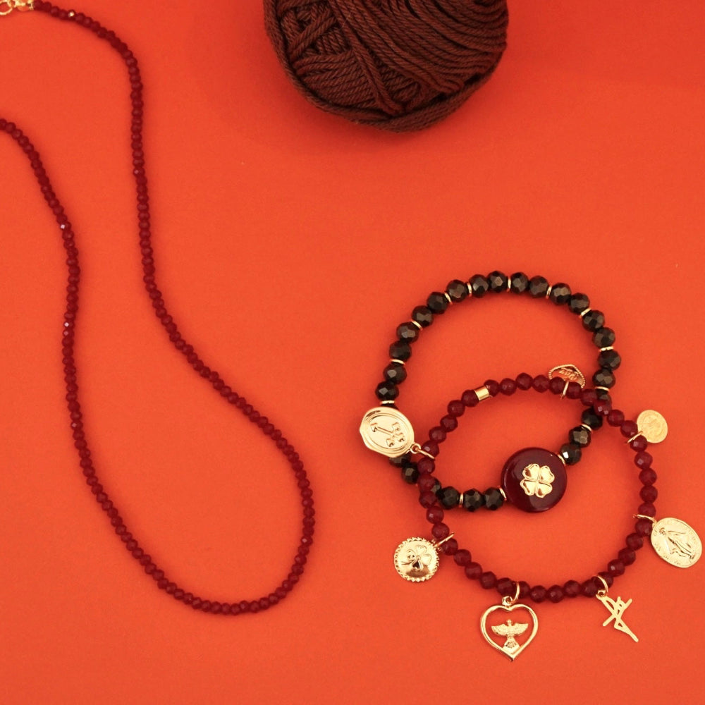 A set of beaded jewelry displayed on a bright orange-red background, including a long dark red bead strand necklace with gold-tone clasp, a stack of three beaded bracelets in dark red, black, and maroon with gold-tone charms, and a ball of dark brown yarn at the top.