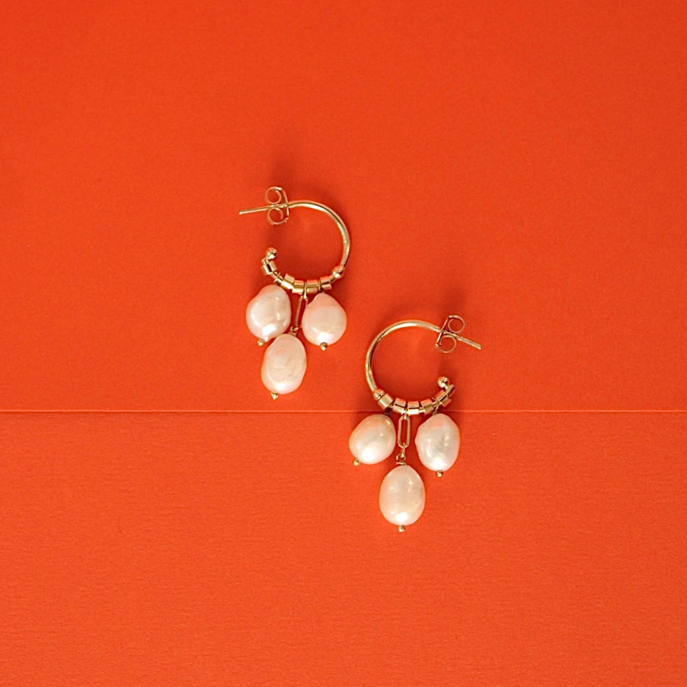 A pair of gold hoop earrings with dangling fresh water pearl, displayed on a solid orange background.