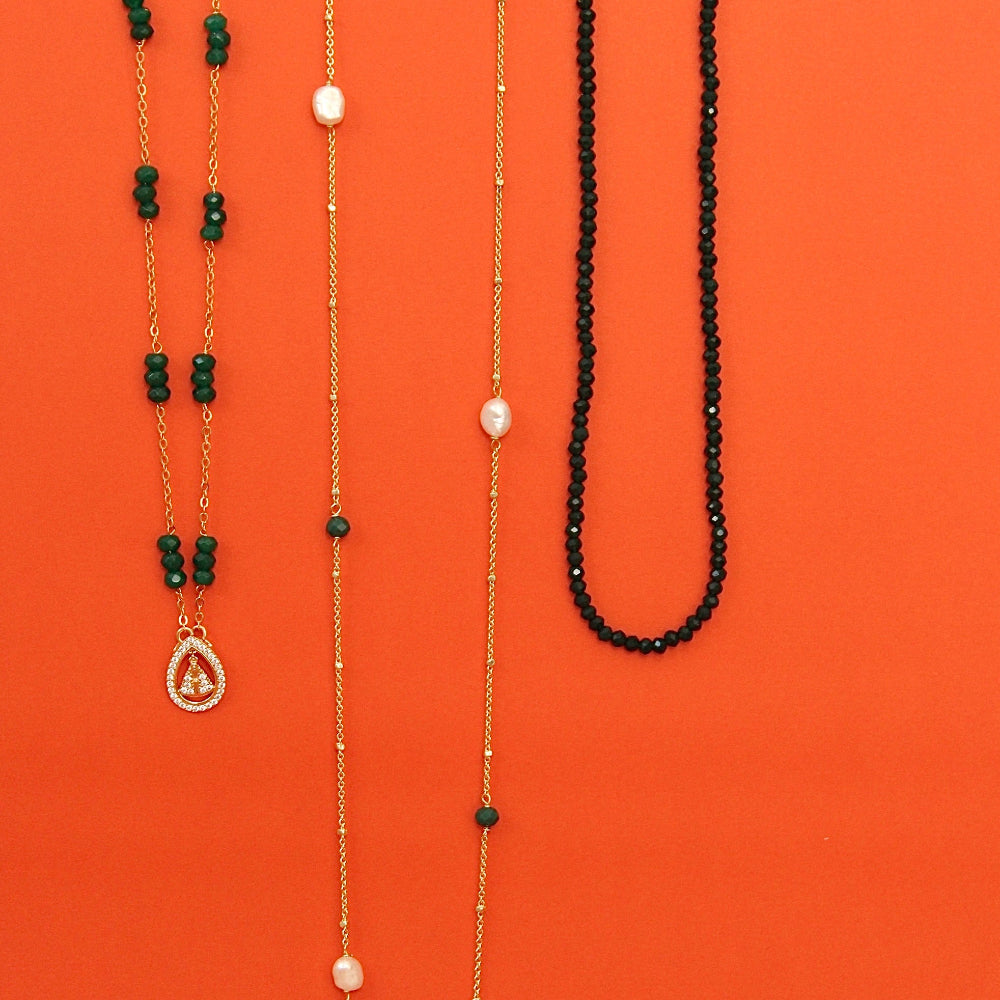 Gold plated necklaces with natural stones displayed vertically against a solid orange background. The left necklace features dark green faceted beads and a teardrop pendant with clear stones.