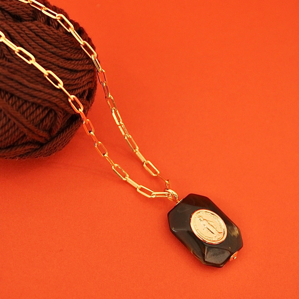 A gold plated chain necklace with a faceted brown pendant featuring a gold plated virgem Mary medal, styled on a vibrant orange background next to a ball of dark brown yarn.