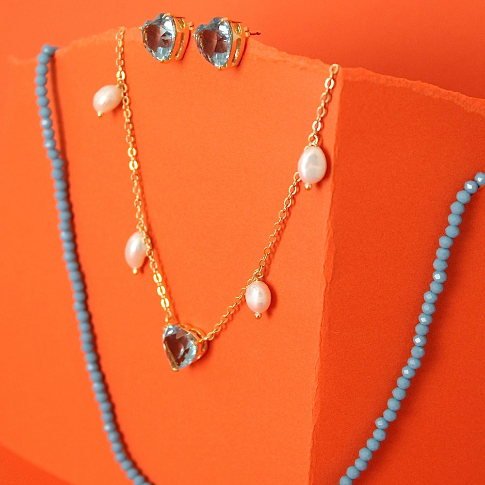 Gold jewelry set with a heart-shaped pendant necklace, matching stud earrings, and a light blue beaded strand draped over a bright orange display block.