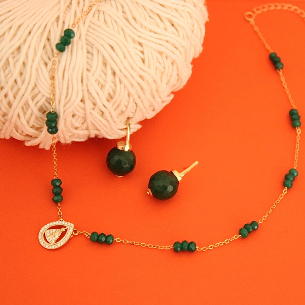 A gold-tone beaded necklace and matching stud earrings with dark green faceted beads, displayed on a bright orange background next to a ball of cream-colored yarn.