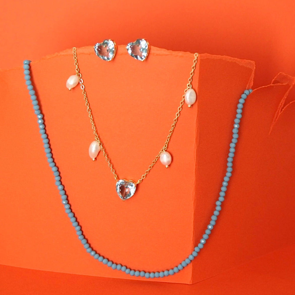 A jewelry set displayed on an orange stand against an orange background, featuring a light blue beaded necklace, a gold chain necklace with pearl-like drops and a heart-shaped clear stone pendant, and matching clear stone stud earrings.
