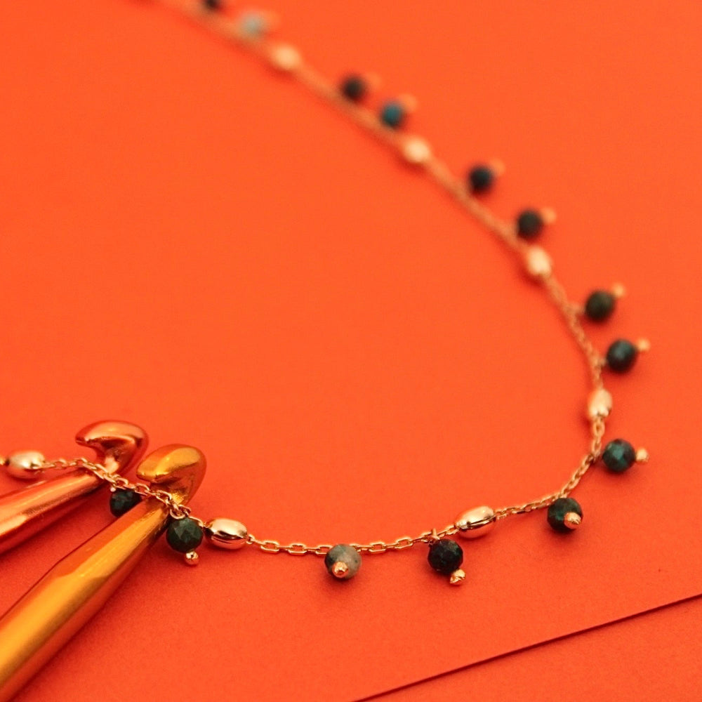 A delicate gold chain necklace with natural stones and gold spacer beads, styled with two gold crochet hooks on a vibrant orange background.
