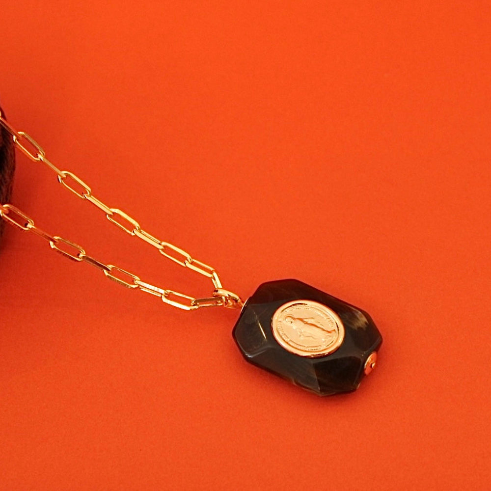 A gold paperclip chain necklace with a faceted black pendant featuring a gold circular inset, displayed on a bright orange background next to a ball of dark brown yarn.