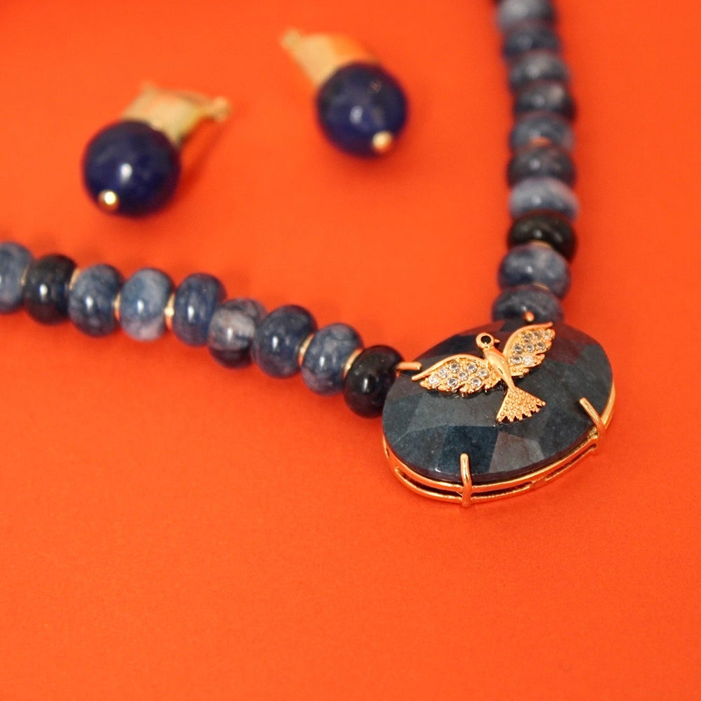 A beaded necklace with dark blue jade natural stones, featuring a central oval dark blue jade stone pendant with a gold bird charm, shown alongside matching blue beads and earrings on a bright orange background.