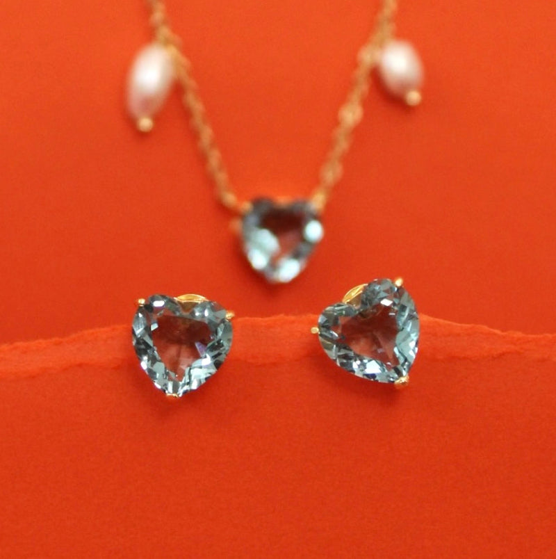 A gold necklace with pearl-like drops and a heart-shaped clear gemstone pendant, displayed above matching heart-shaped stud earrings on a bright orange background.