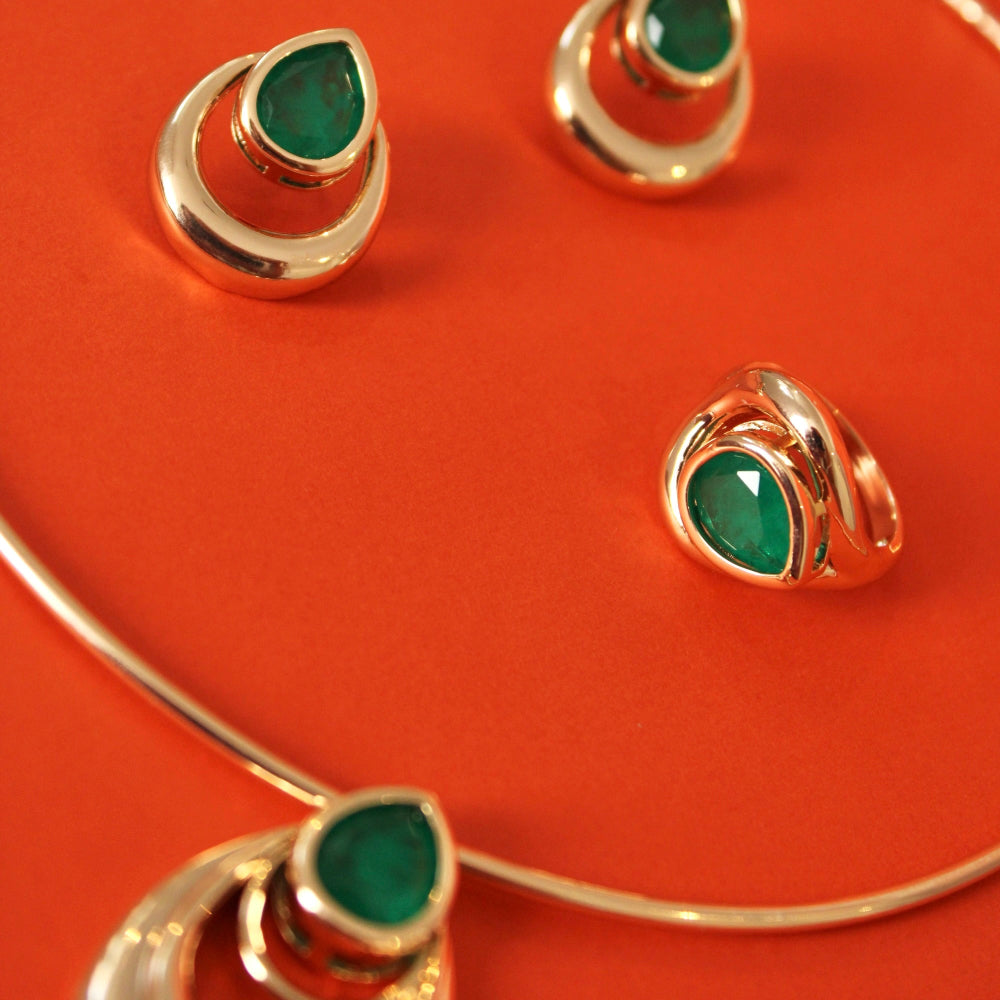 A gold jewelry set featuring a necklace, earrings, and ring, each adorned with a teardrop-shaped green emerald gemstone, displayed on a vibrant orange-red background.