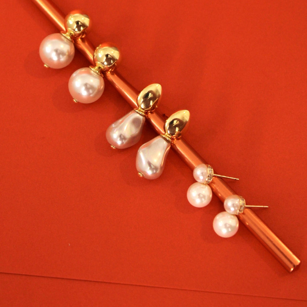 A gold-toned crochet hook decorated with dangling pearl beads and small pearl stud earrings, displayed diagonally on a solid red background.