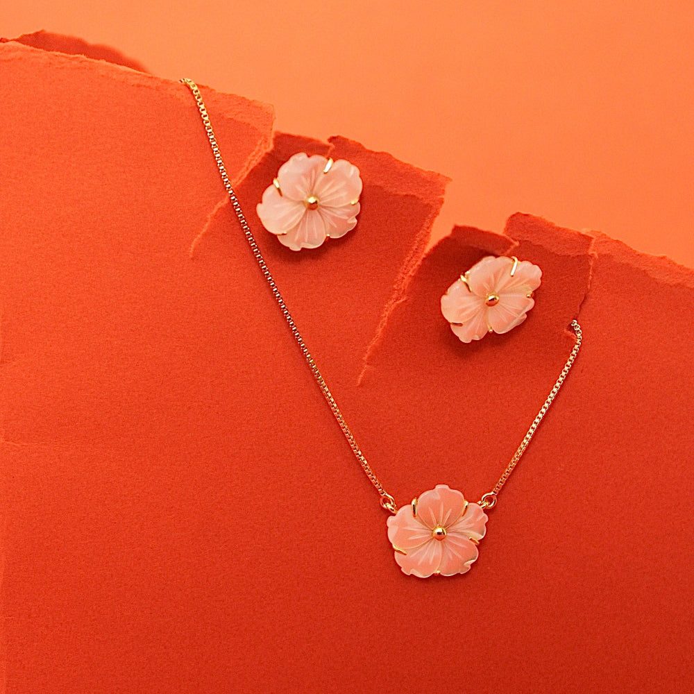 A gold-tone necklace and matching stud earrings set with pink cherry blossom flower pendants, displayed on torn orange paper against a solid orange background.