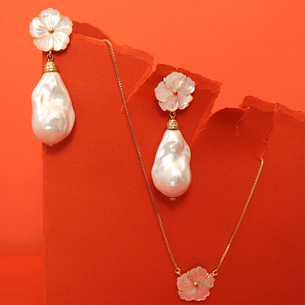 A jewelry set featuring a gold necklace and matching drop earrings, each adorned with a white baroque pearl and a mother of pearl flower-shaped charm, displayed against a torn orange-red background.