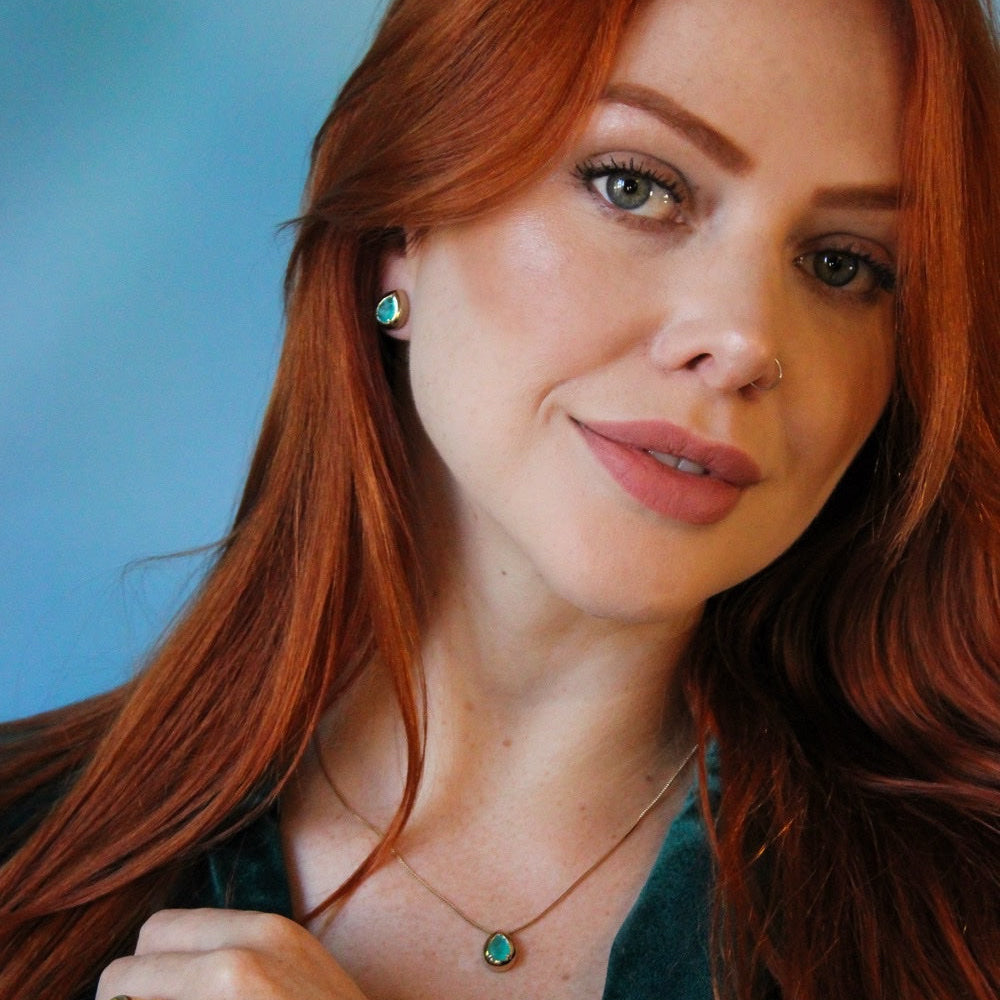 A close-up portrait of a woman with long red hair wearing a gold necklace and matching stud earring with oval green stones, set against a solid blue background.