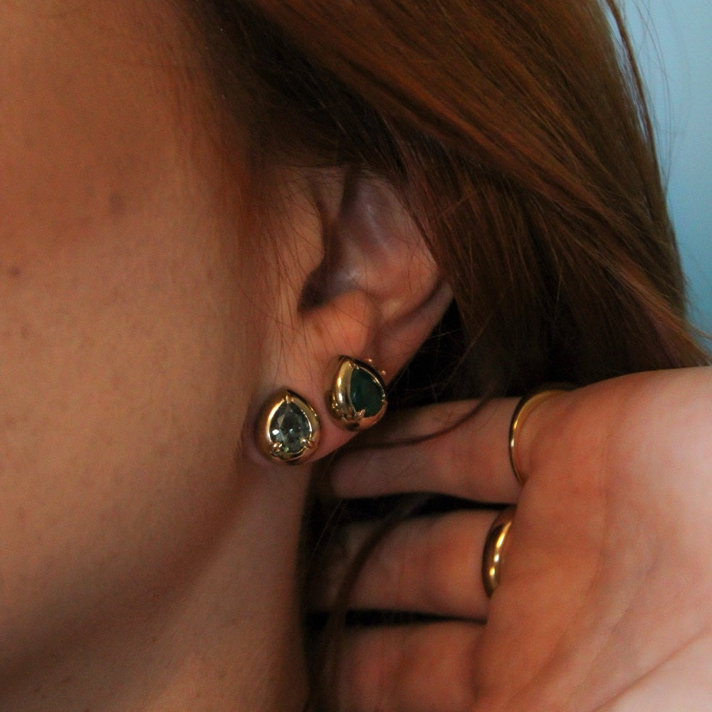 Close-up of a person wearing gold stud earrings with oval green stones, with their hand near their ear.