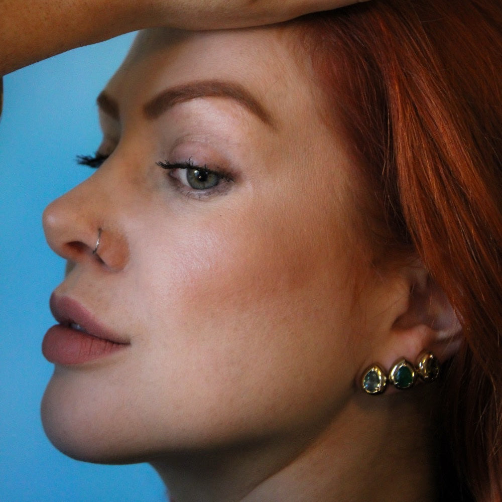 A close-up portrait of a woman with red hair wearing a large gold ring with three green stones and matching green stone earrings, with her hand resting on her forehead against a solid blue background.