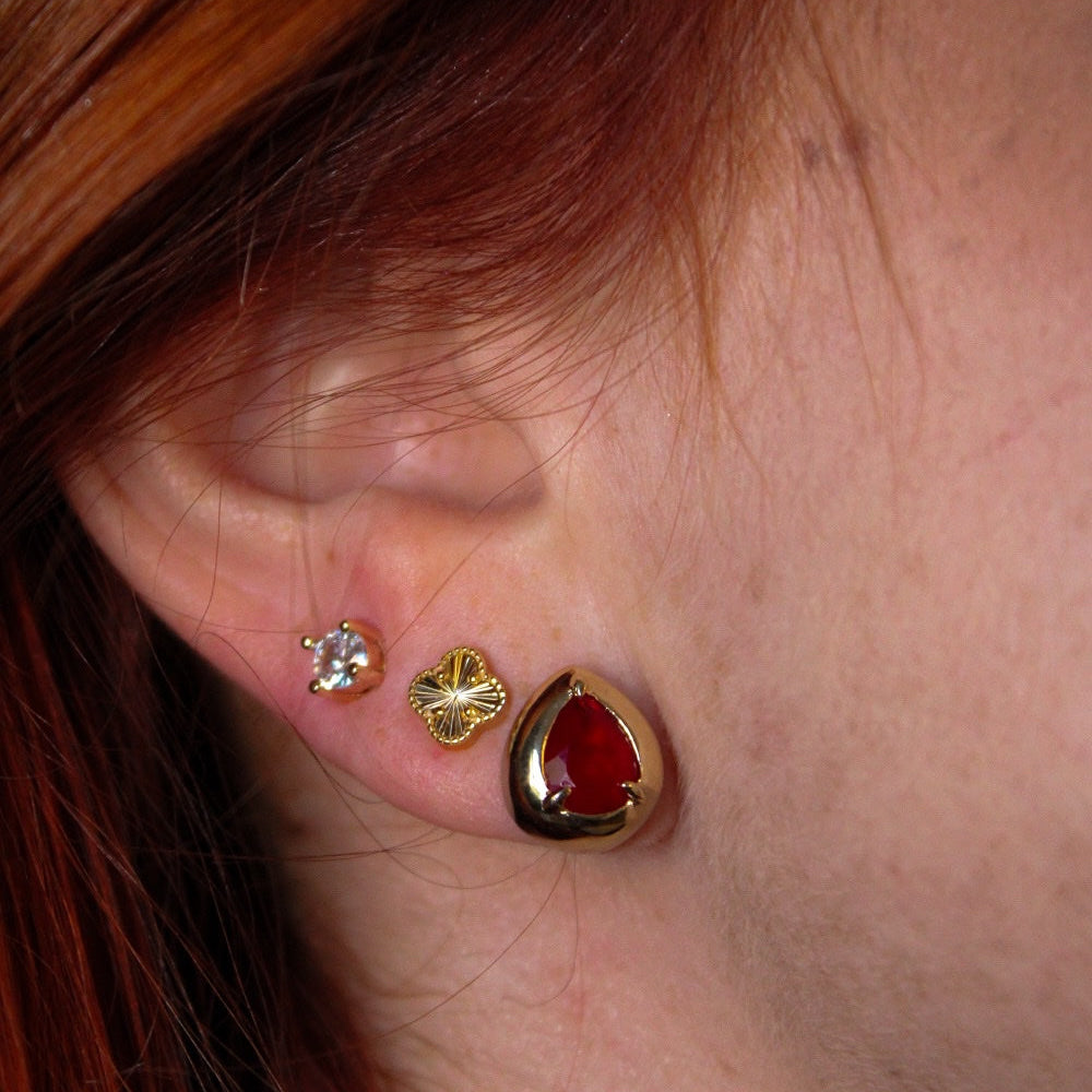 Close-up of a person's ear wearing three gold-toned earrings: a small January birthstone gem stud, a gold clover stud, and a larger gold teardrop stud with a red heart-shaped center