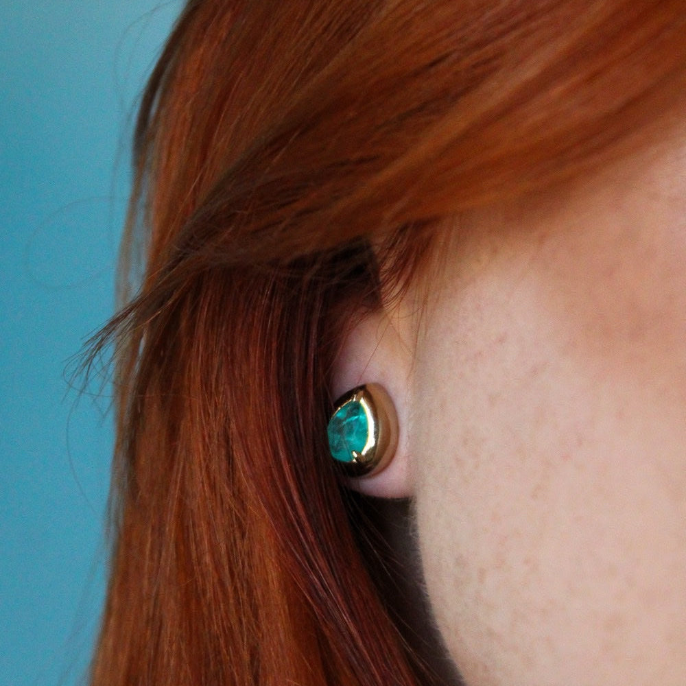 A close-up of a person's ear wearing a gold-toned stud earring with a marquise-cut turquoise gemstone, set against a solid blue background.