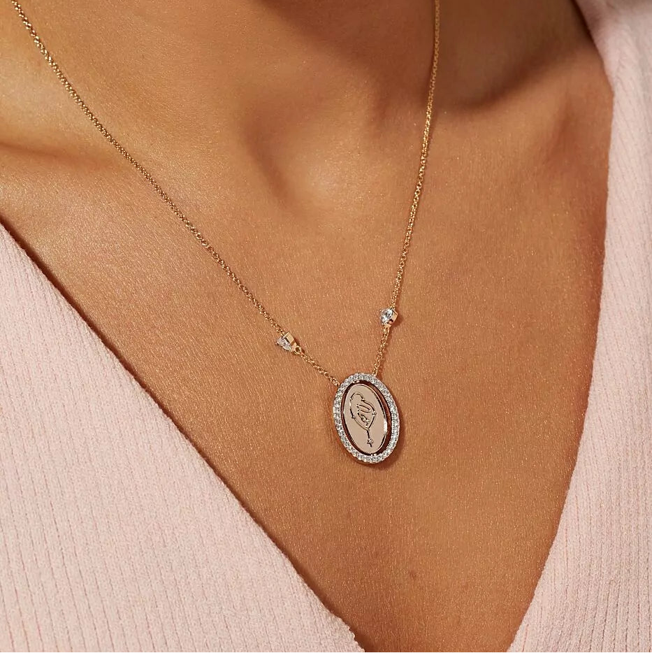 A close-up of a woman wearing a delicate gold chain necklace with an oval pendant featuring a floral engraving, set against a soft pink top.