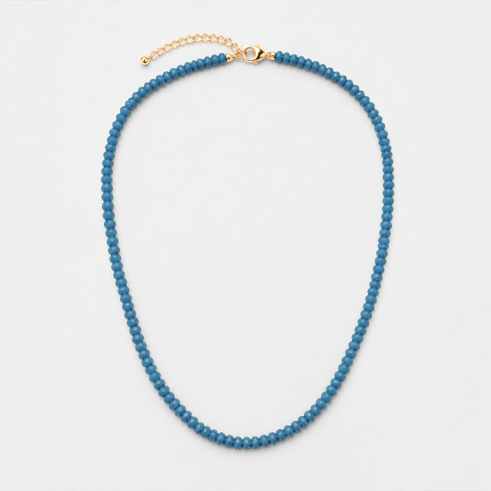 A beaded necklace made of small, round blue beads with a gold-tone clasp and extender chain, displayed on a clean white background.