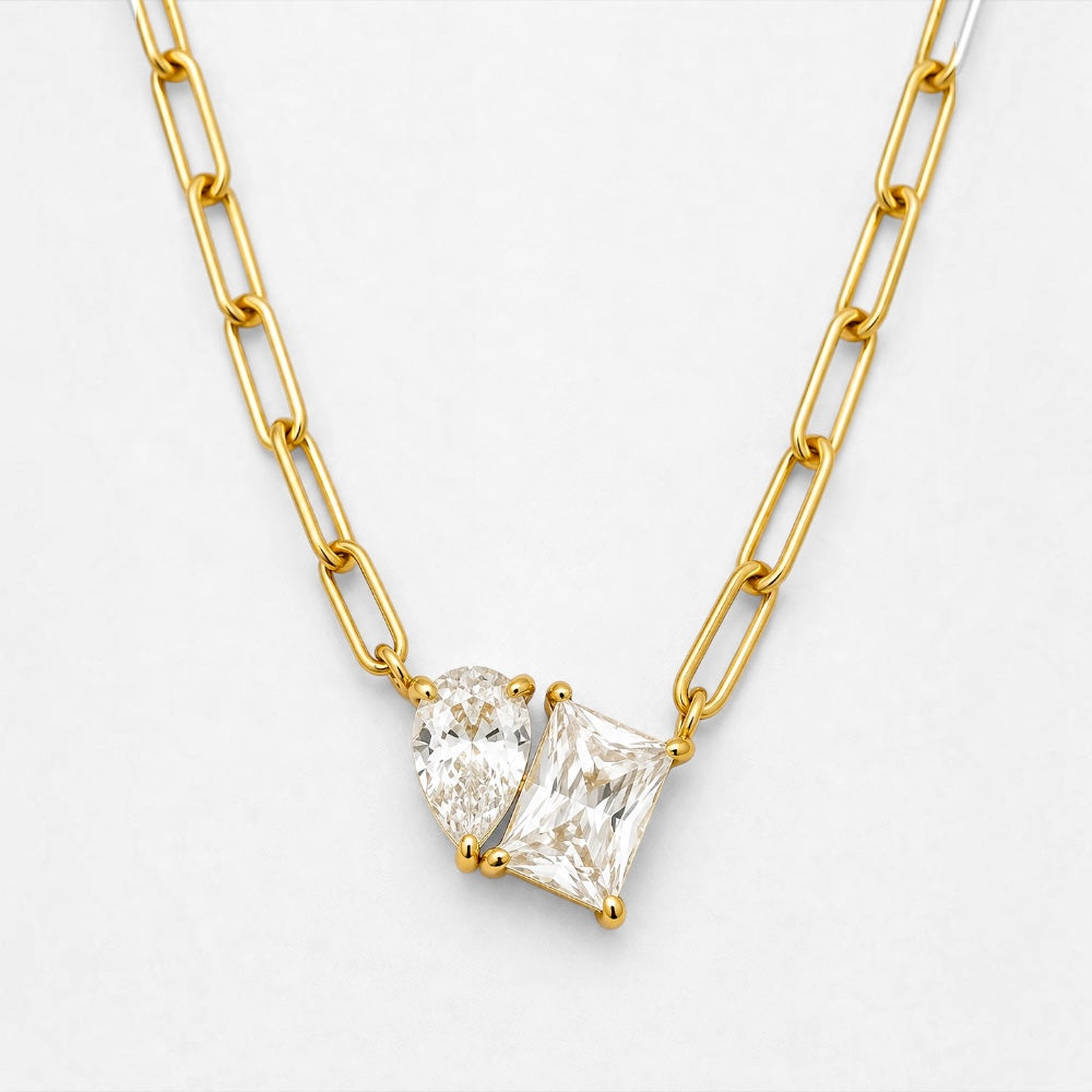 A gold necklace featuring a dual-stone pendant with a pear-cut and rectangular princess-cut clear gemstone, set on a gold paperclip chain against a light gray background.