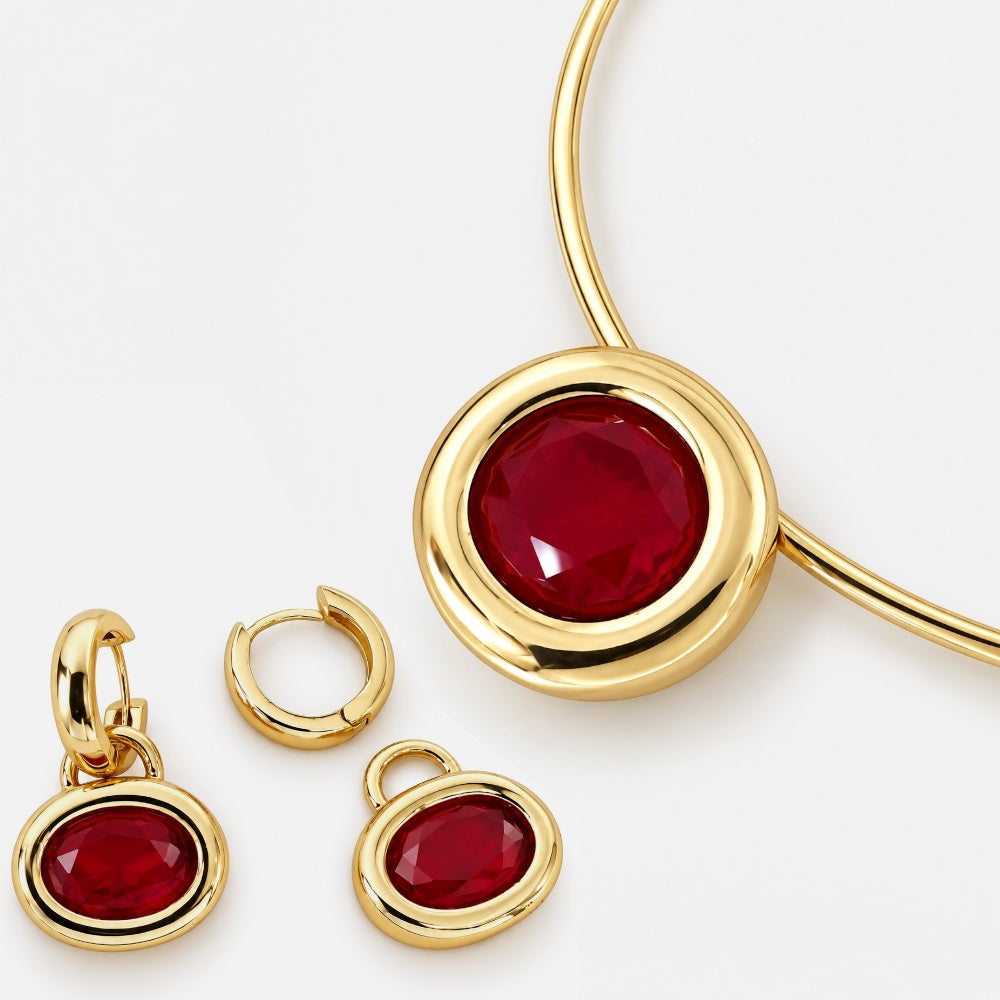 Gold jewelry set featuring a round bezel-set red gemstone pendant on a bangle, a matching small oval drop earring, and a matching hoop earring, shown on a clean white background.