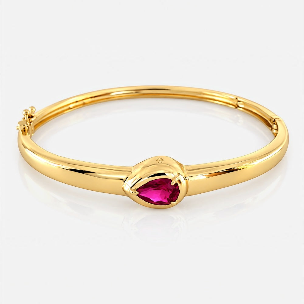 A polished yellow gold bangle bracelet featuring a central teardrop-shaped pink gemstone, set against a clean white background with a soft reflection.