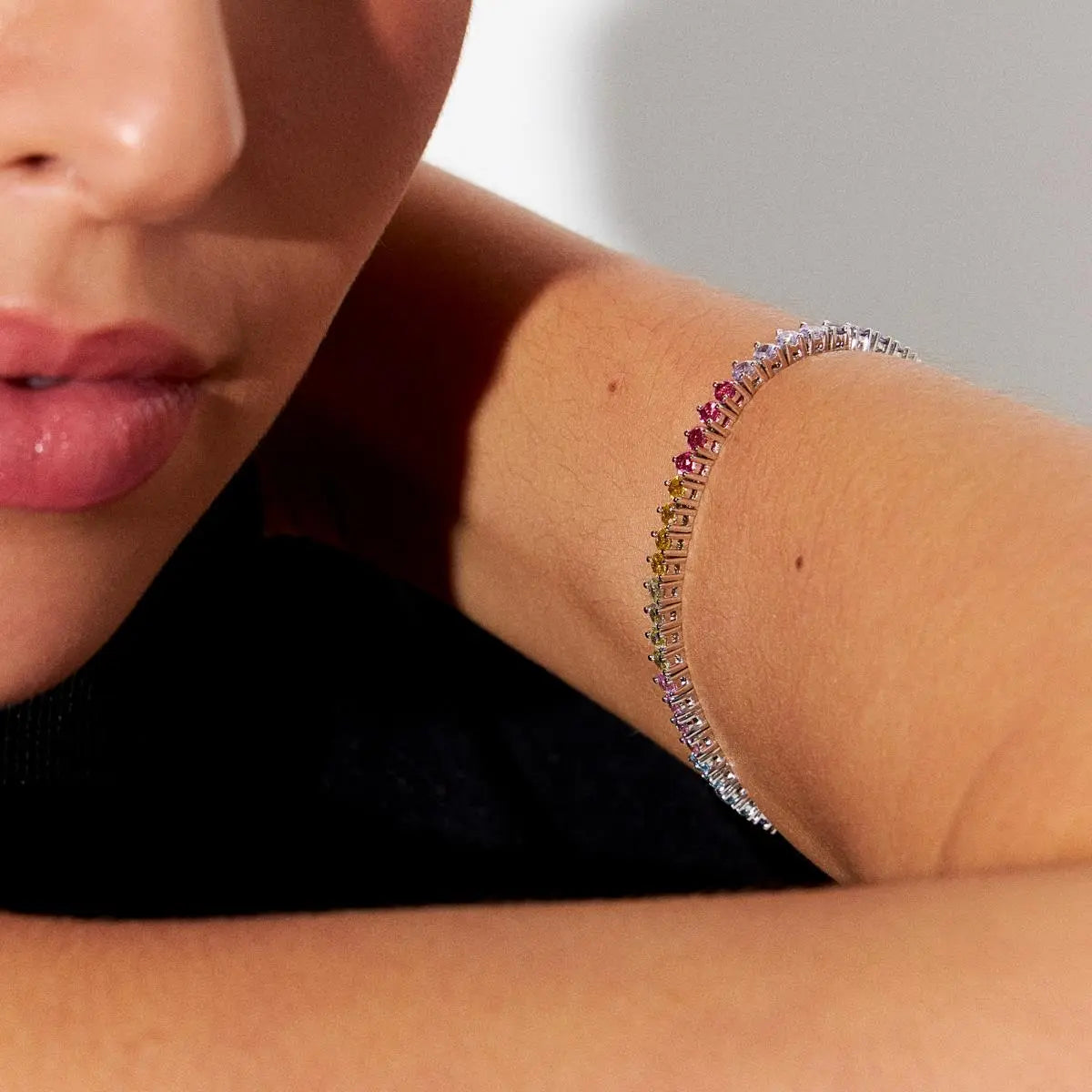 A close-up lifestyle shot of a model's wrist wearing a delicate silver tennis bracelet adorned with small, colorful square-cut gemstones.