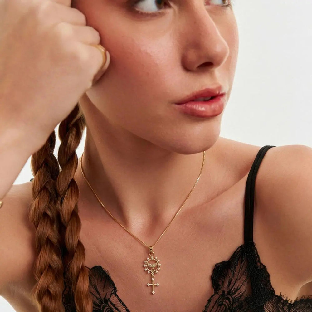 A close-up lifestyle shot of a woman wearing a delicate gold key pendant necklace with a small cross at the bottom, styled with a black lace camisole against a clean white background.