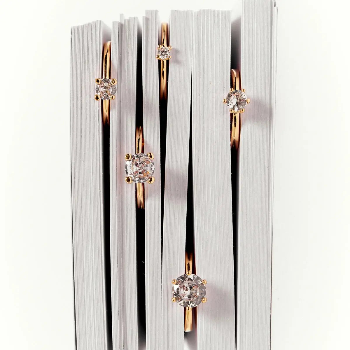 Four rose gold bookmarks with clear round gemstones, arranged vertically between the pages of a white book.