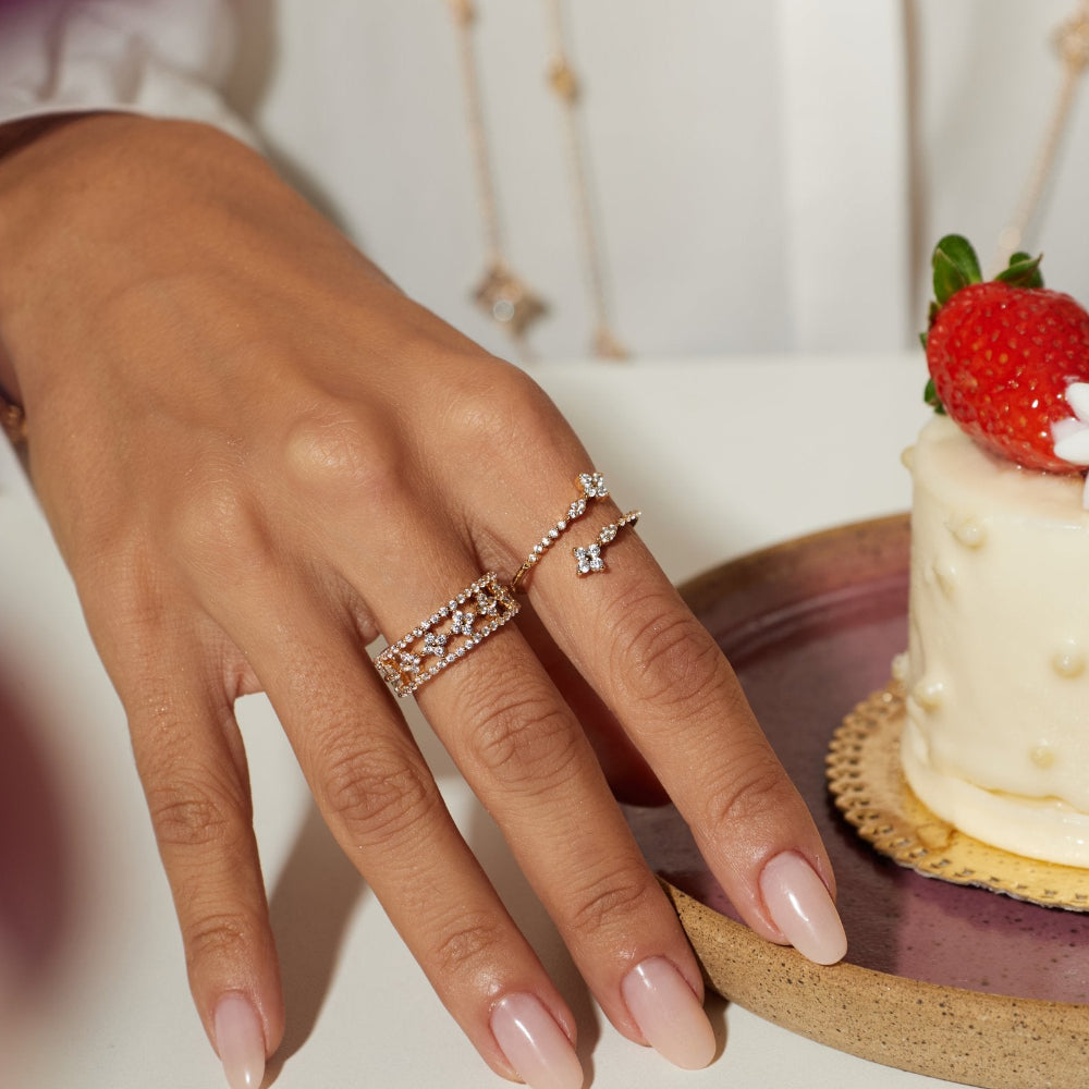 A close-up of a person's hand resting on a white surface next to a small cream-colored dessert topped with a strawberry, wearing two delicate silver rings with small clear stones.