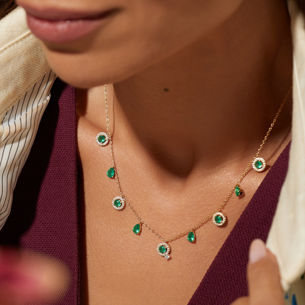 Close-up of a model wearing a delicate gold necklace with green emerald-cut stones and a matching ring, styled with a deep burgundy top.